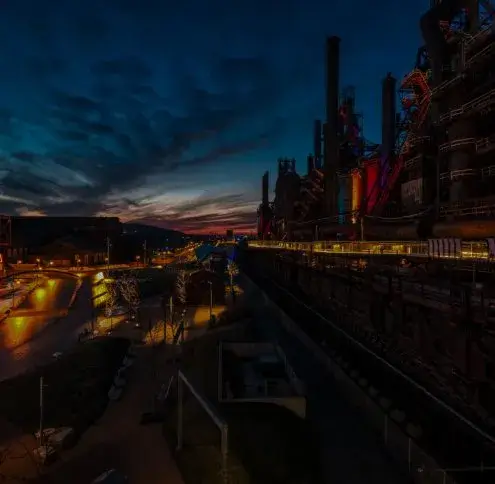 Lehigh Valley service area: floodlit blast furnaces and smokestacks of a steel plant standing above a lit street at dusk